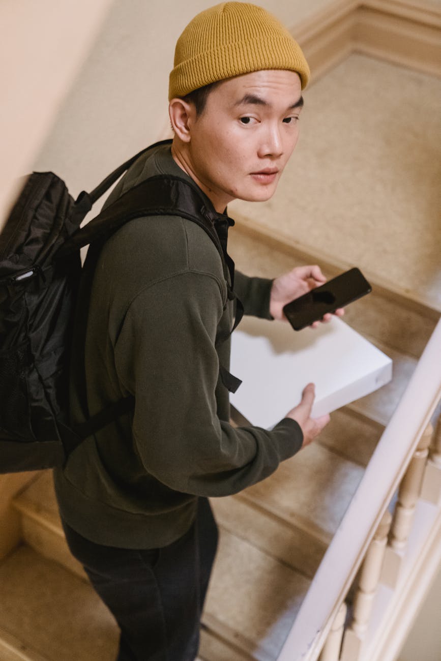 Young man with a backpack and beanie delivering a package on stairwell, holding a phone.