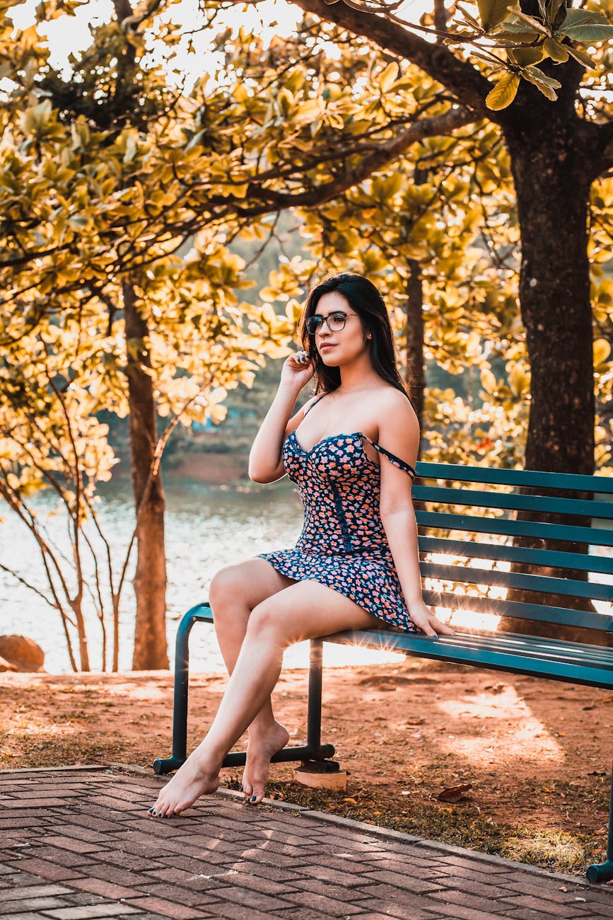 A young woman in a floral dress sitting on a bench in a sunny park, surrounded by trees.