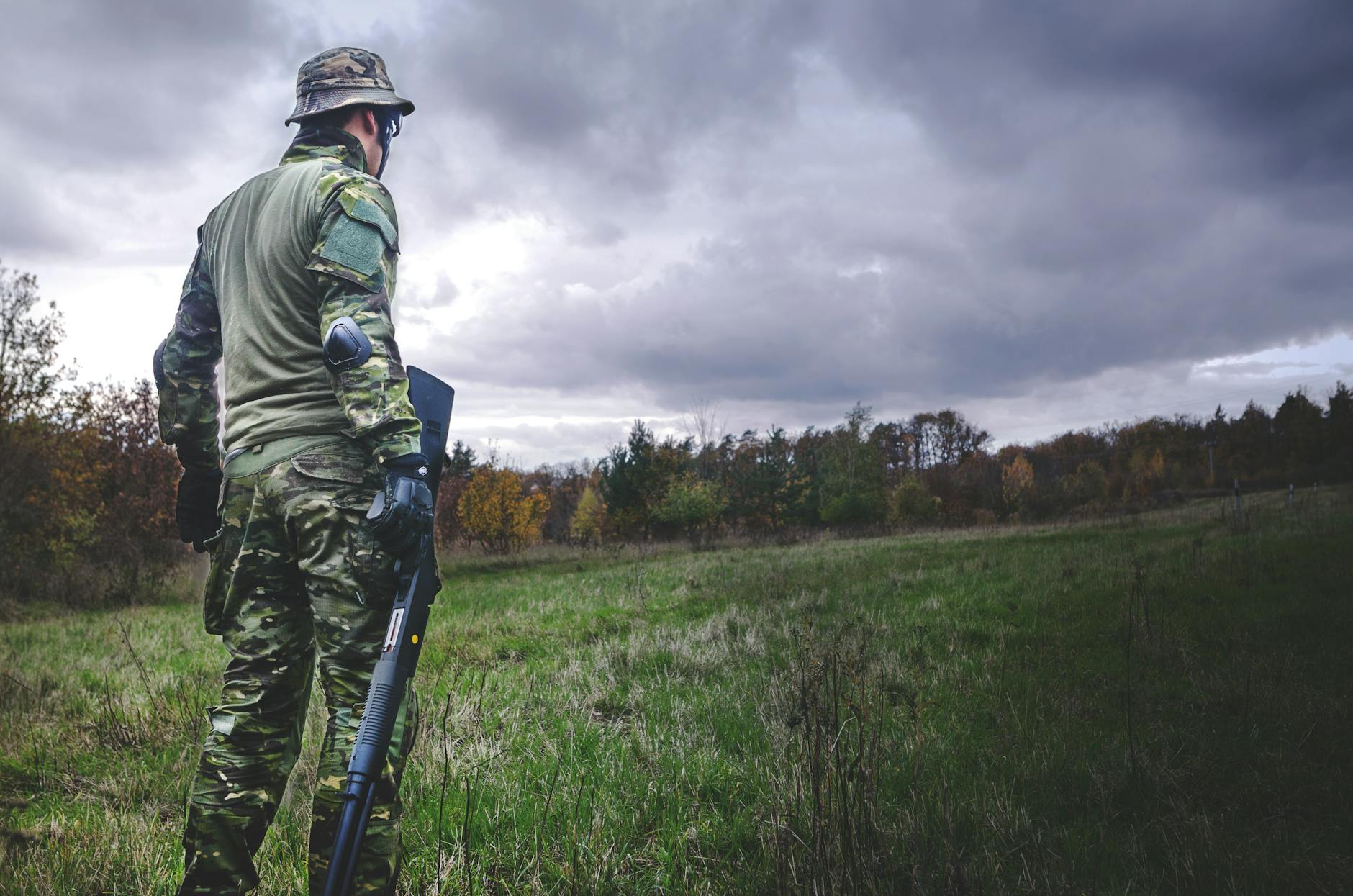 A soldier in camouflage with a rifle stands in a grassy field under a cloudy sky.