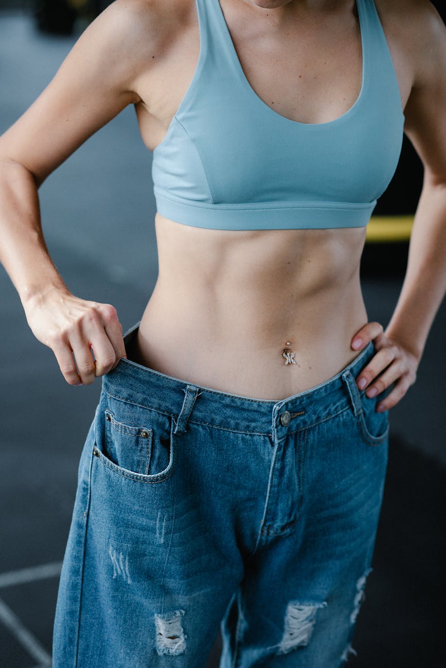 Fit woman shows weight loss in oversized jeans, displaying toned abs.