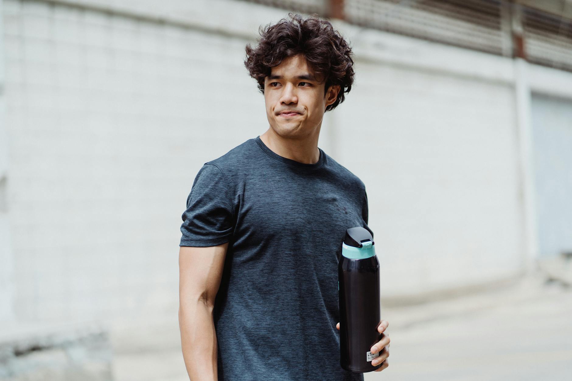 Casual young man with curly hair holding a water bottle outdoors on a bright day.