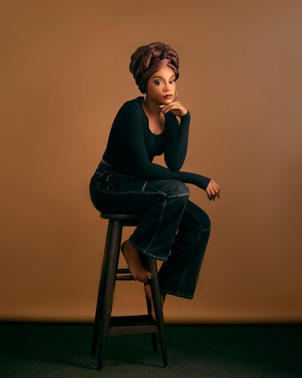 Stylish woman in headwrap and jeans sitting on a stool with a warm background.