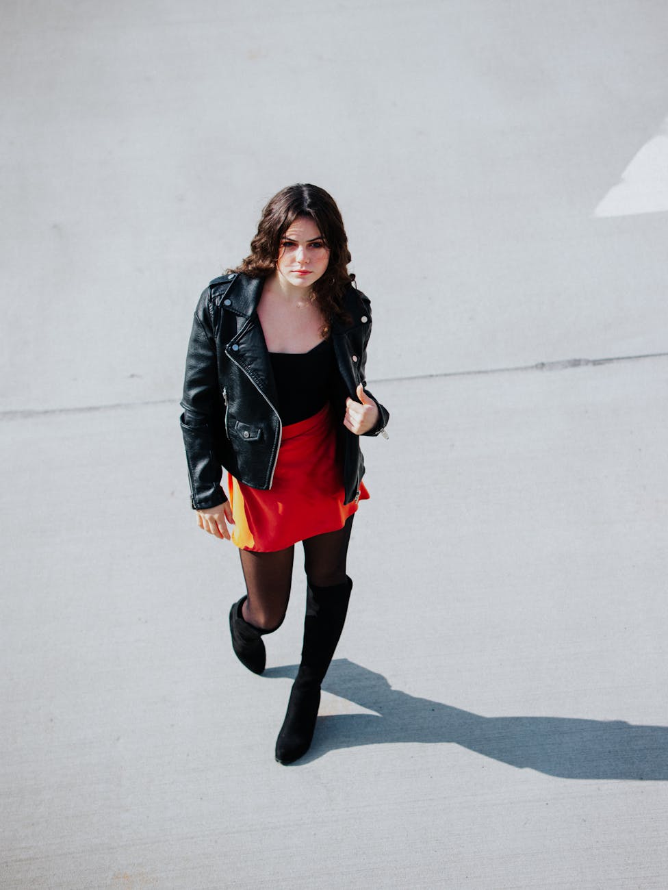Chic young woman in red skirt and black jacket walking outdoors in bright daylight.
