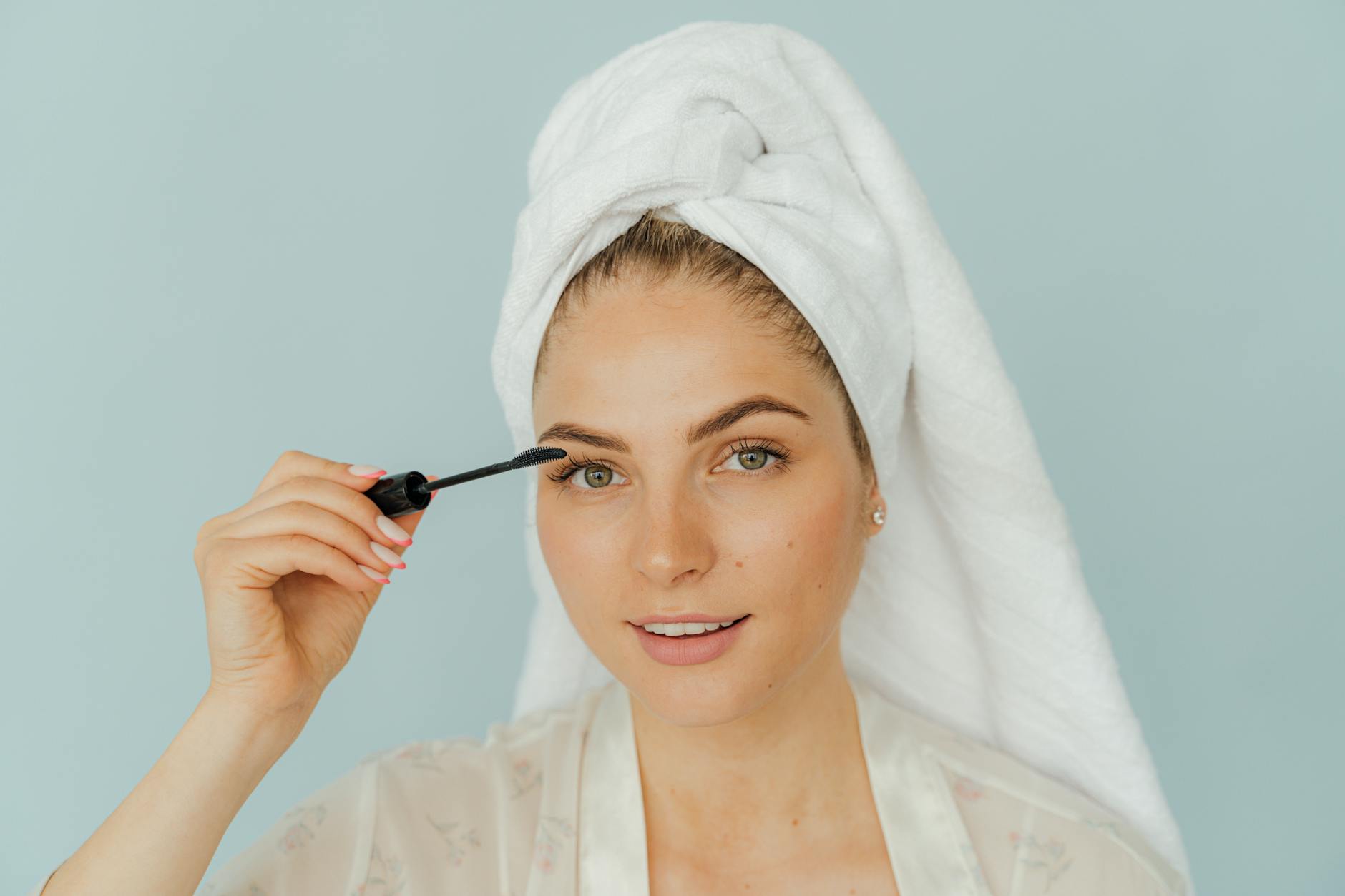 A woman smiles while applying mascara, wrapped in a towel headwrap against a light blue background.