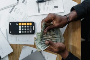 Person counting dollar bills over documents with a smartphone calculator on the desk.